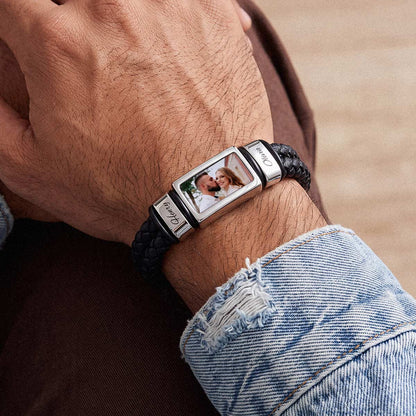 A close-up of a custom photo bracelet worn on a person's wrist. The bracelet features a black braided leather band with a polished silver-toned rectangular metal centerpiece.