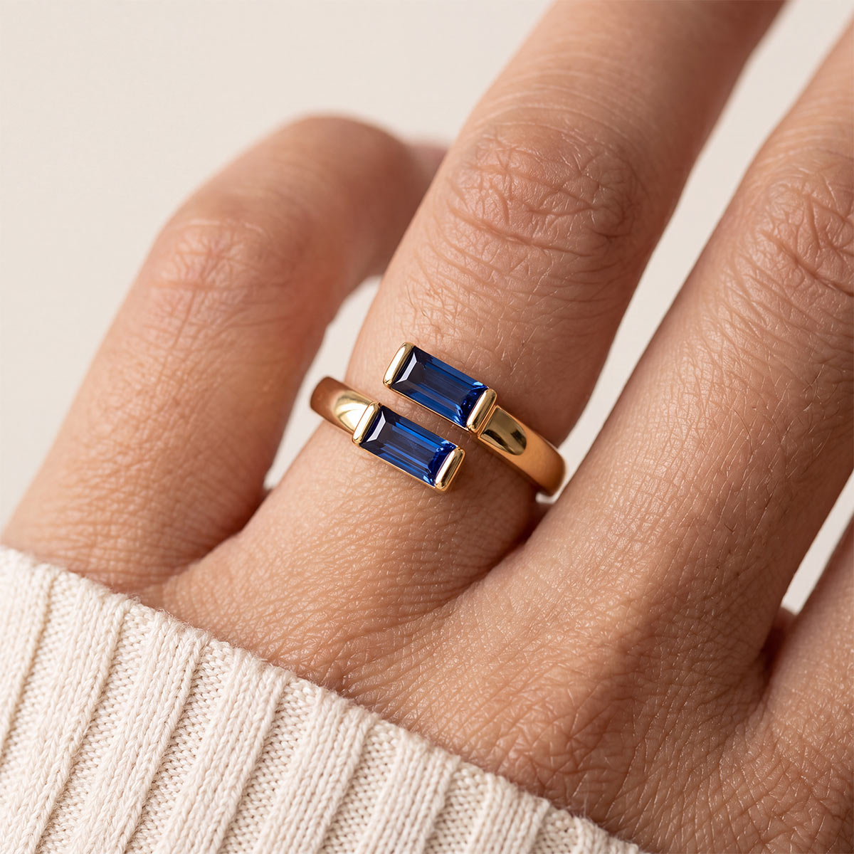 A close-up of a gold birthstone ring with two rectangular blue gemstones worn on a finger, set against a soft beige background.