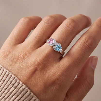A close-up of a hand wearing a silver birthstone infinity ring with a heart-shaped pink gemstone and a blue heart-shaped gemstone, accented by small clear stones.