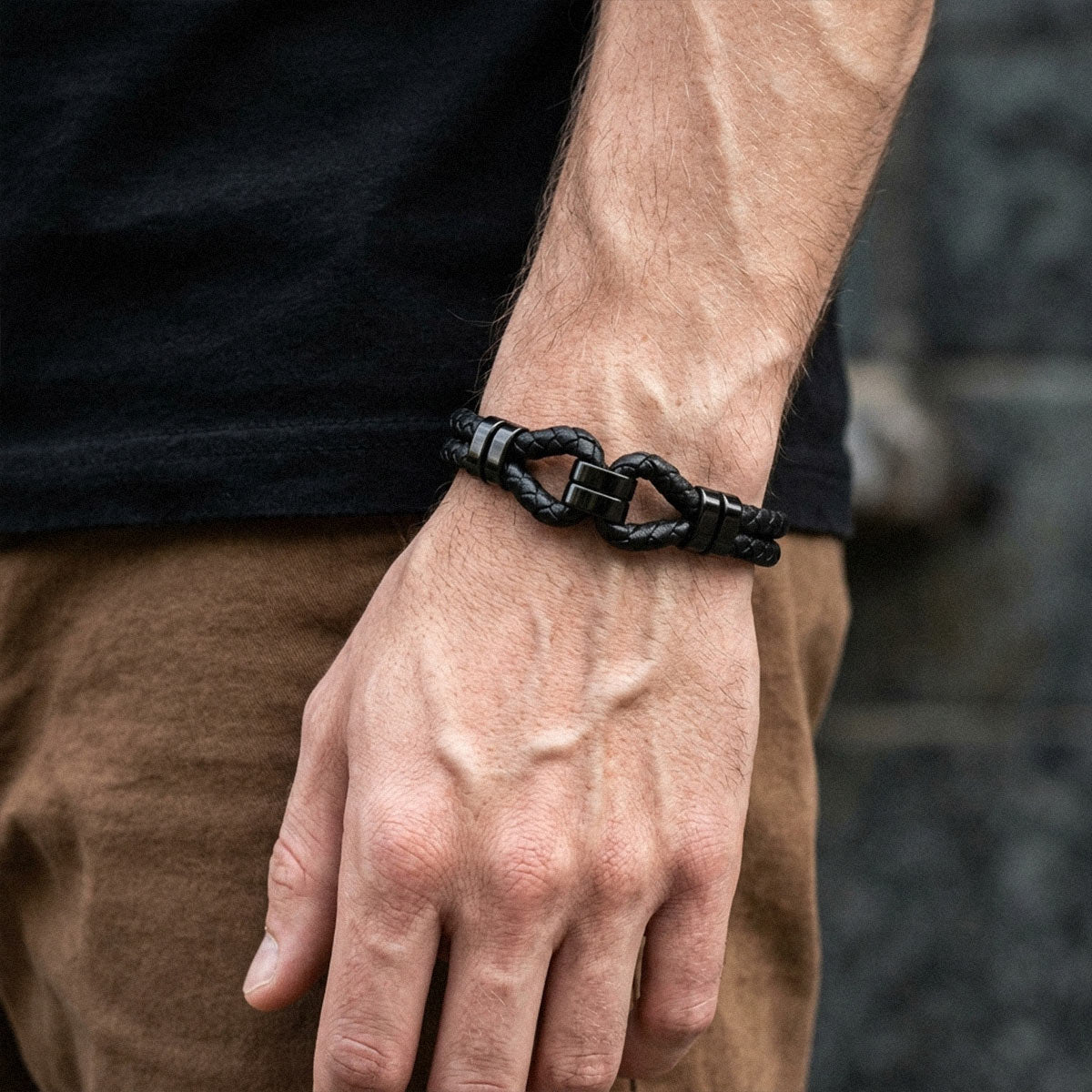 A close-up of a man's hand and wrist wearing a black braided cord bracelet with a large interlocking link clasp, paired with a black t-shirt and brown pants.