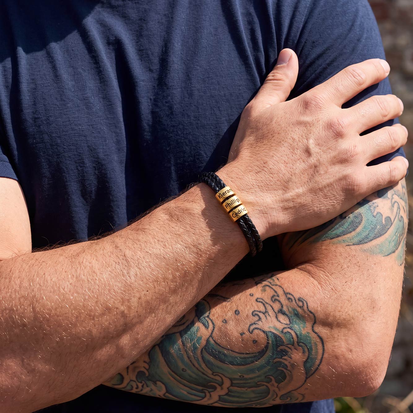 A close-up of a man's tattooed arm wearing a black braided bracelet with three gold cylindrical beads, set against a navy blue shirt.