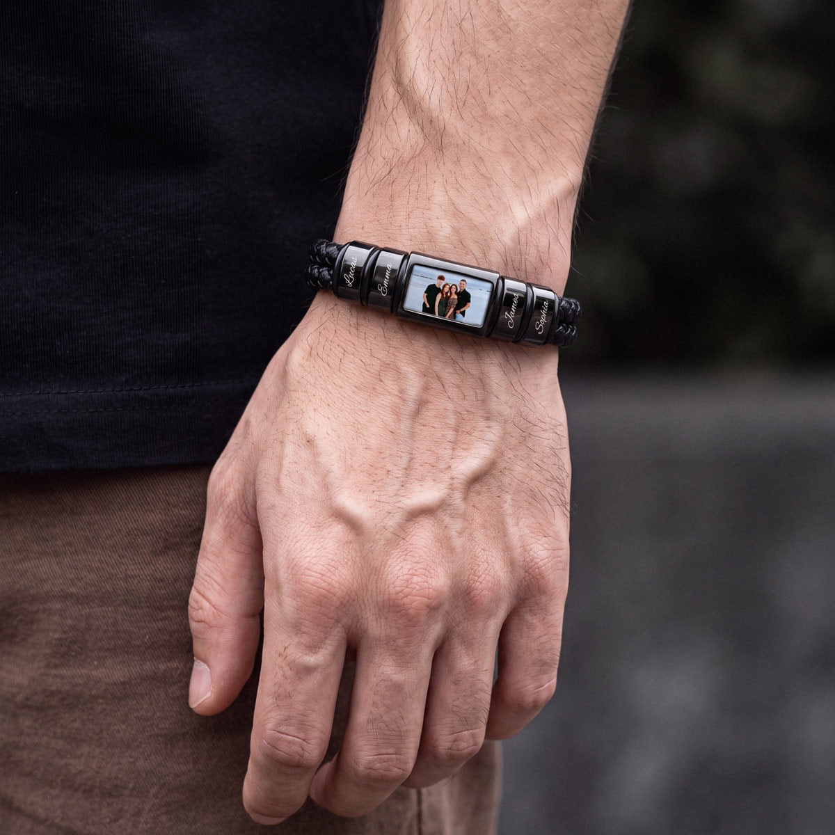 A close-up of a man's wrist wearing a black beaded bracelet with a central rectangular compartment displaying a custom photo of two people.