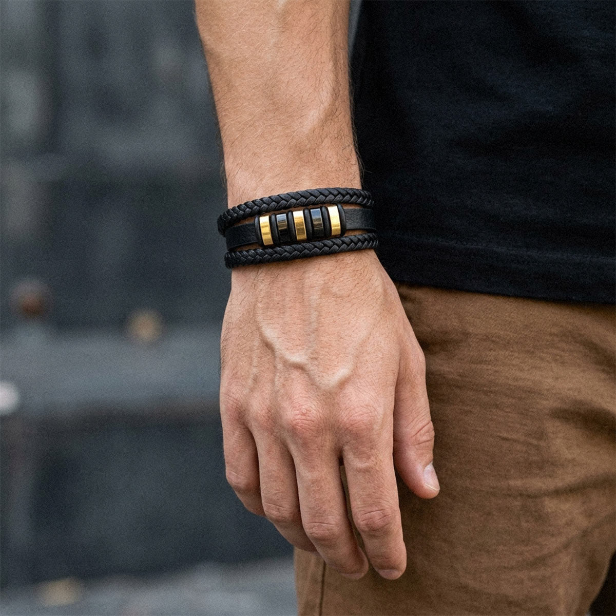 A close-up of a man's wrist wearing a black braided leather bracelet with three gold-toned cylindrical accents, paired with a black shirt and brown pants.