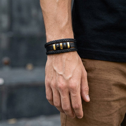 A close-up of a man's wrist wearing a black braided leather bracelet with three gold-toned cylindrical accents, paired with a black shirt and brown pants.