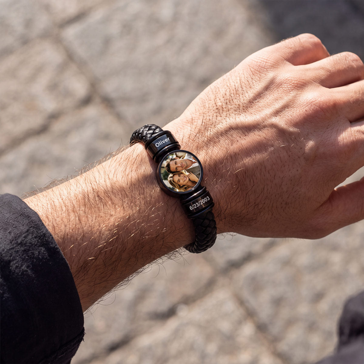 A close-up of a man's wrist wearing a black braided leather photo bracelet with a custom circular dial photo featuring a couple, set against a blurred stone pavement background.
