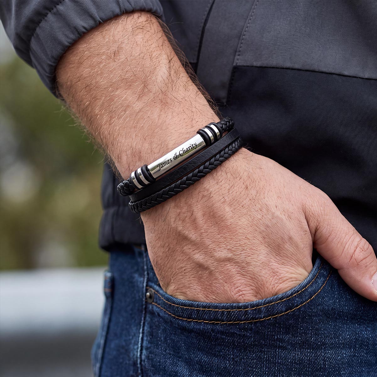 A close-up of a man's wrist wearing a black leather bracelet with a polished silver central bar engraved with 'Proud Father of Chelsea' and black-and-white striped accents, paired with dark gray and blue clothing.