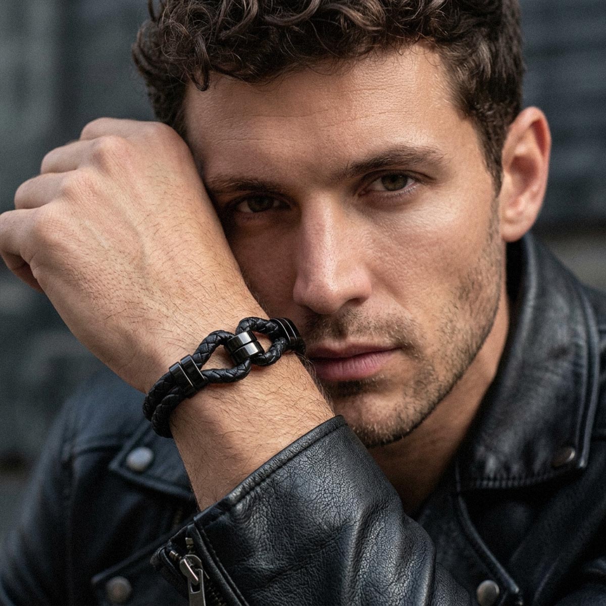 A close-up of a man wearing a black braided leather bracelet with a silver-toned metal clasp, resting his head on his hand in a leather jacket.