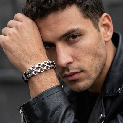 A close-up of a man wearing a chunky silver chain-link bracelet on his wrist, dressed in a black leather jacket against a dark gray background.