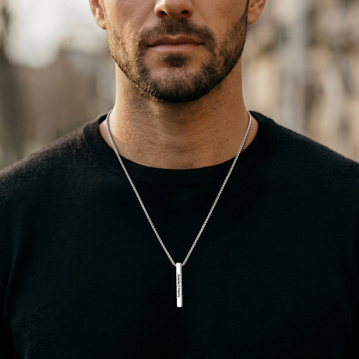 A close-up of a man wearing a silver vertical engraved bar pendant necklace with engraved text, styled with a black crewneck sweater against a blurred outdoor background.