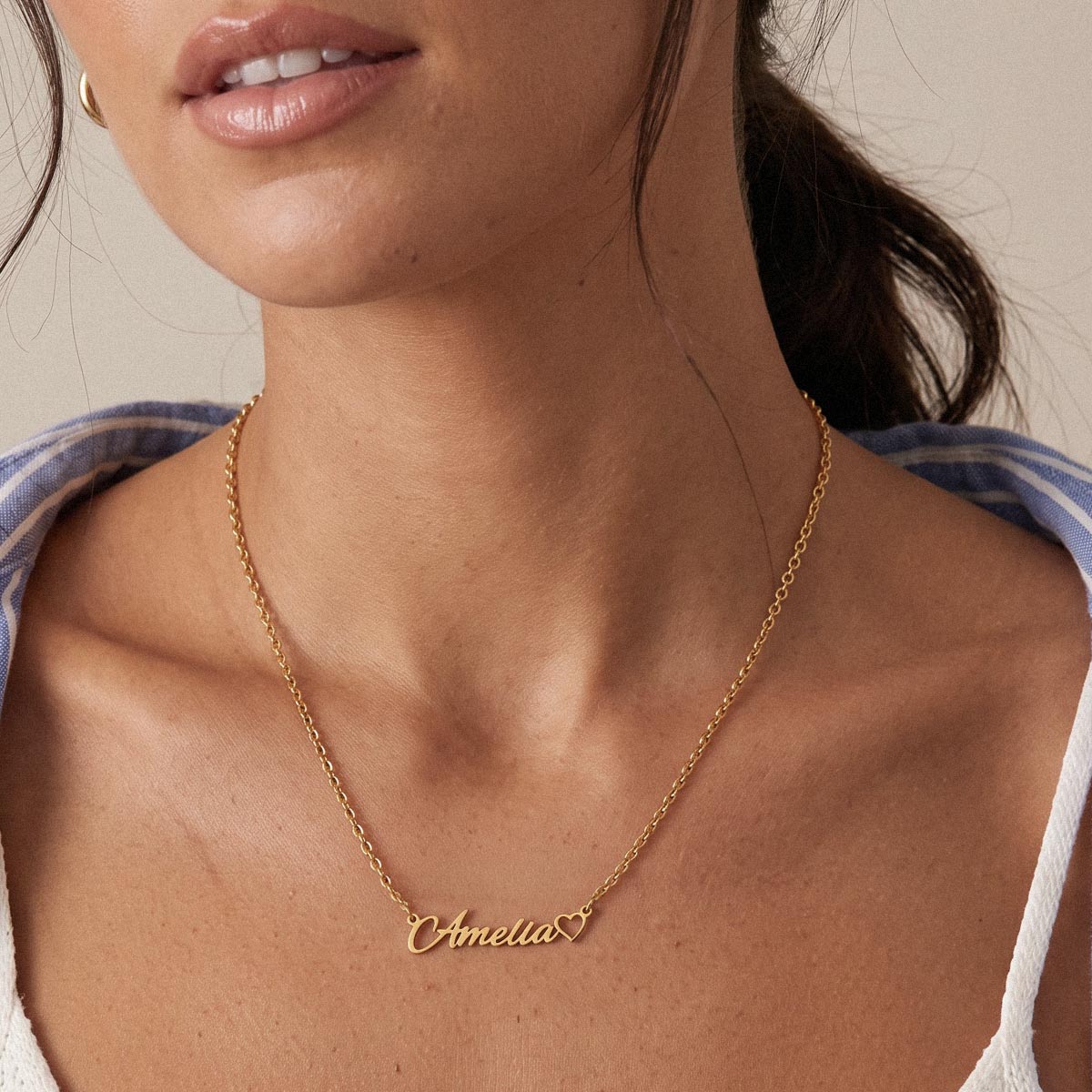 A close-up of a model wearing a delicate gold name necklace with a custom script nameplate reading 'Amelia' and a small heart at the end.