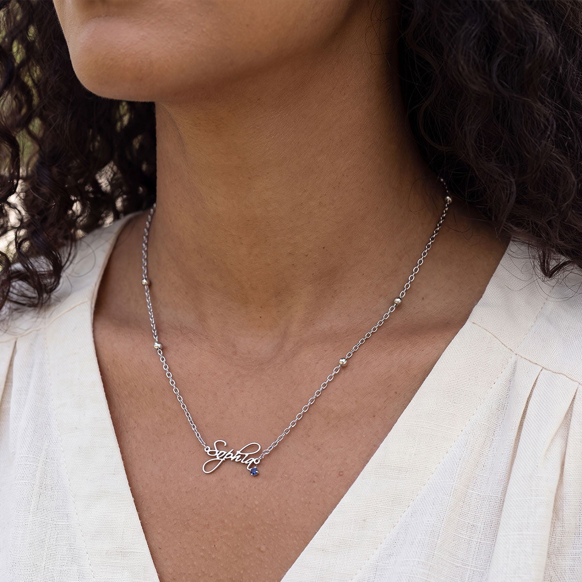 A close-up of a model wearing a silver custom name necklace with the name 'Sophia' and a small dark blue stone accent.