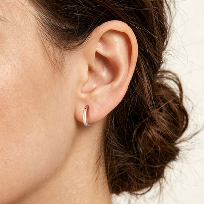 A close-up of a person's ear wearing a small silver huggie earring, with dark brown hair pulled back against a plain light background.