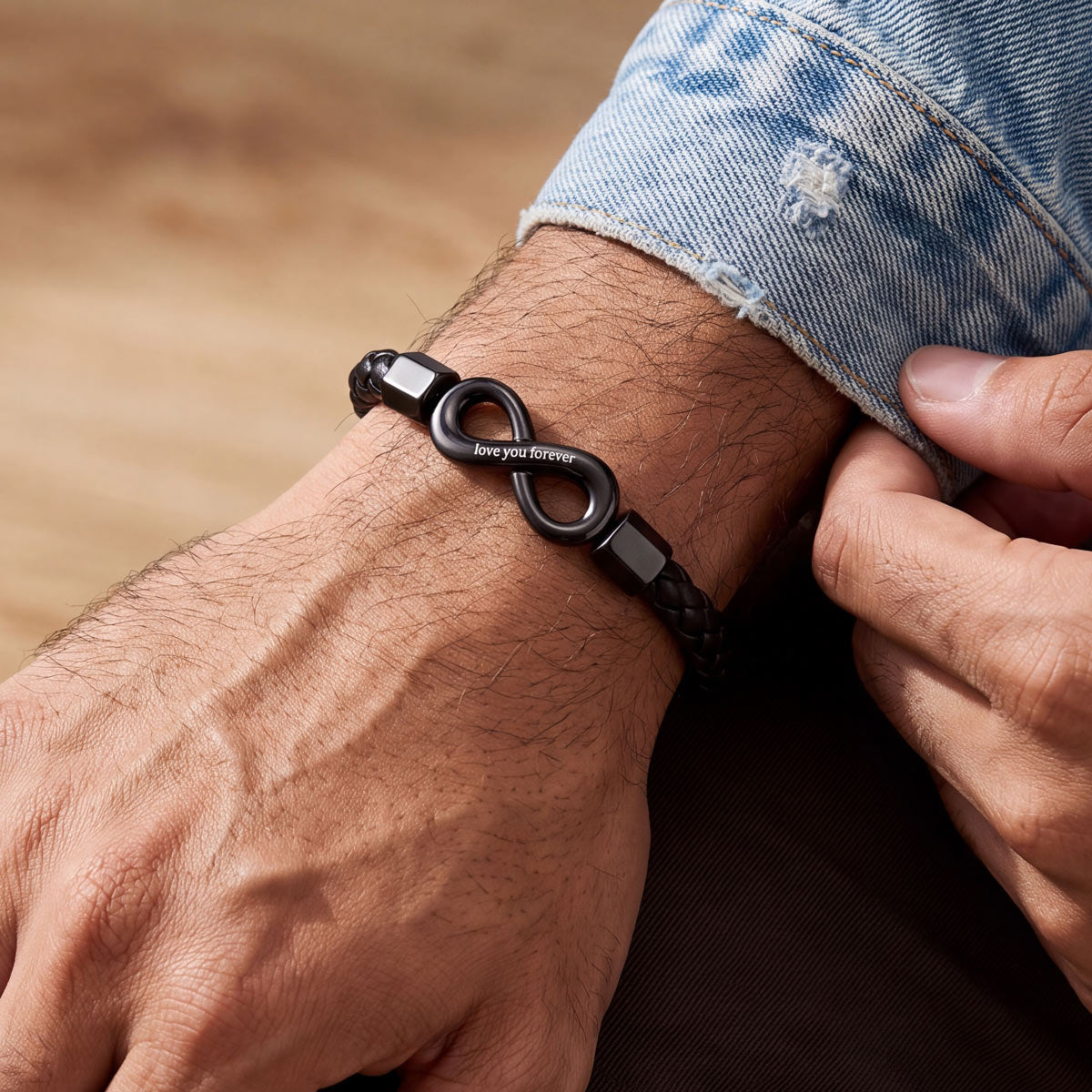 A close-up of a person's wrist wearing a black braided bracelet with a central infinity symbol and small engraved text, paired with a light blue denim cuff.