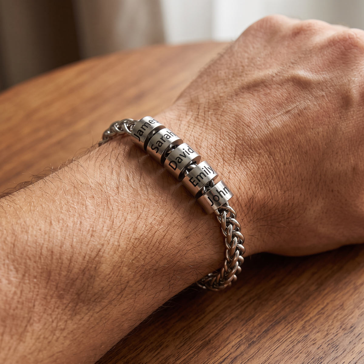 A close-up of a person's wrist wearing a personalized silver name bracelet with round metal beads spelling out the names 'Emily', 'Daniel', 'David', and 'Joseph' on a wooden table.