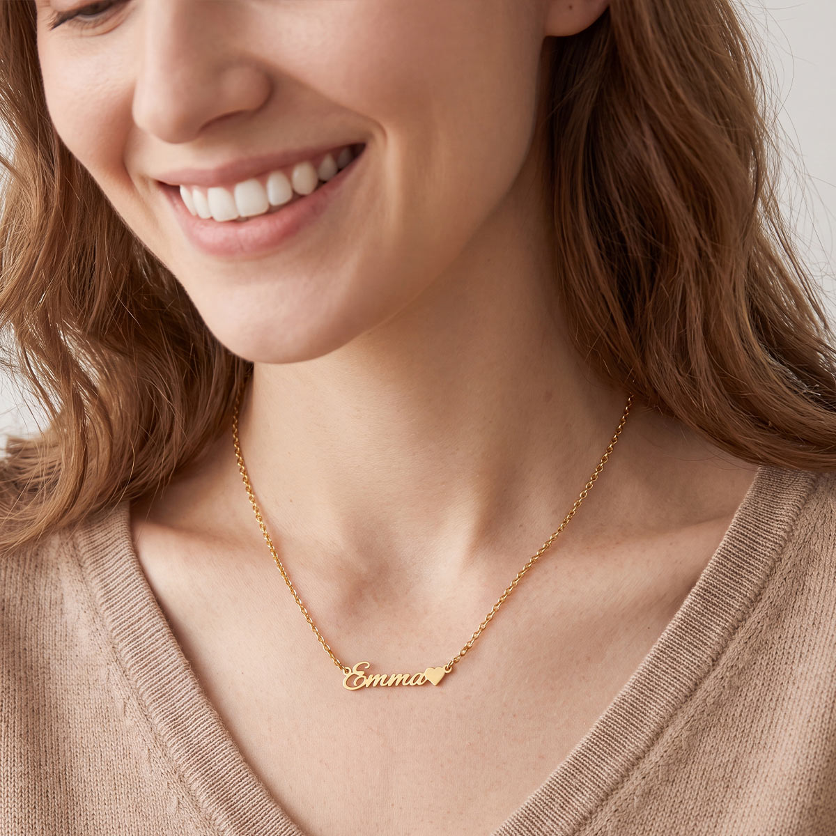 A close-up of a smiling woman wearing a delicate gold chain custom name necklace with the name 'Emma' and a small heart in script on a light gray background.