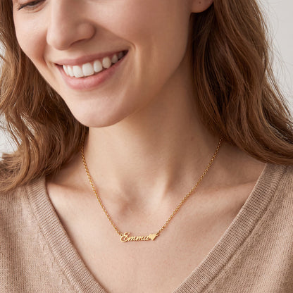 A close-up of a smiling woman wearing a delicate gold chain custom name necklace with the name 'Emma' and a small heart in script on a light gray background.