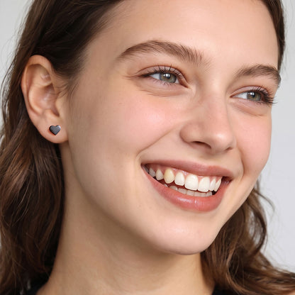 A close-up of a smiling woman wearing a small dark heart-shaped stud earring on her earlobe, set against a clean light gray background.