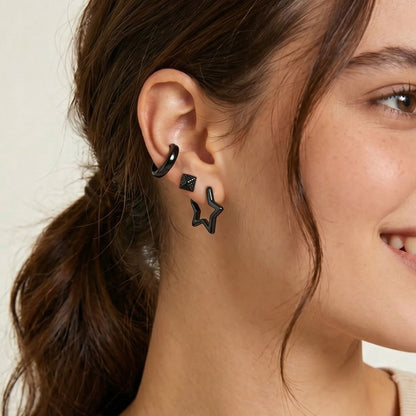 A close-up of a woman's ear wearing three black stud and hoop earrings, including a small diamond-shaped stud, a curved hoop, and two open star-shaped studs.