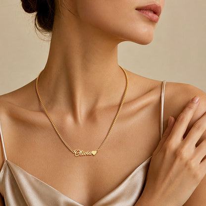 A close-up of a woman wearing a delicate gold chain necklace with a personalized script nameplate reading 'Olivia' and a small heart, styled with a satin camisole against a warm beige background.