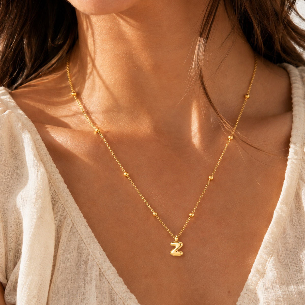 A close-up of a woman wearing a delicate gold initial necklace with small bead accents and a lowercase letter 'z' pendant, styled with a white top.
