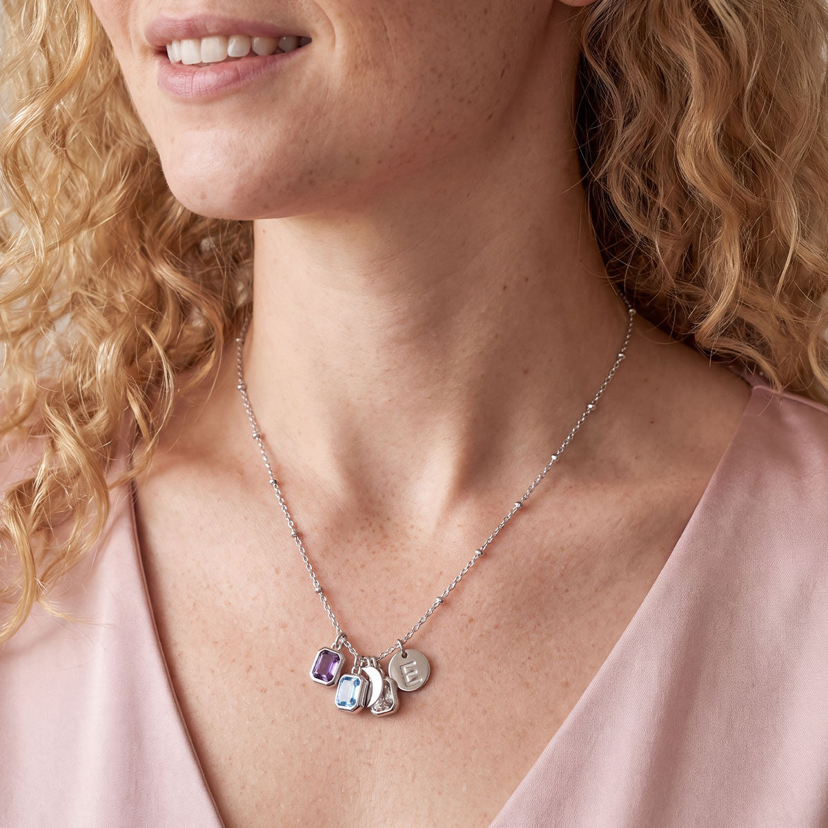 A close-up of a woman wearing a delicate silver intial birthstone necklace with four small charms, including a purple stone, a blue stone, a silver horse, and a silver monogram pendant. She is wearing a soft pink satin top.