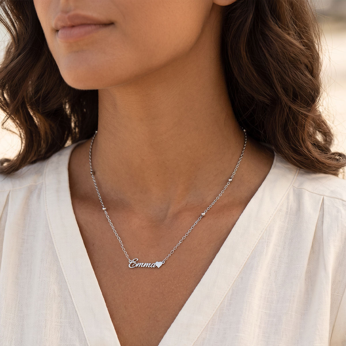 A close-up of a woman wearing a delicate silver nameplate necklace that reads 'Emma' on a fine chain, paired with a white V-neck blouse.