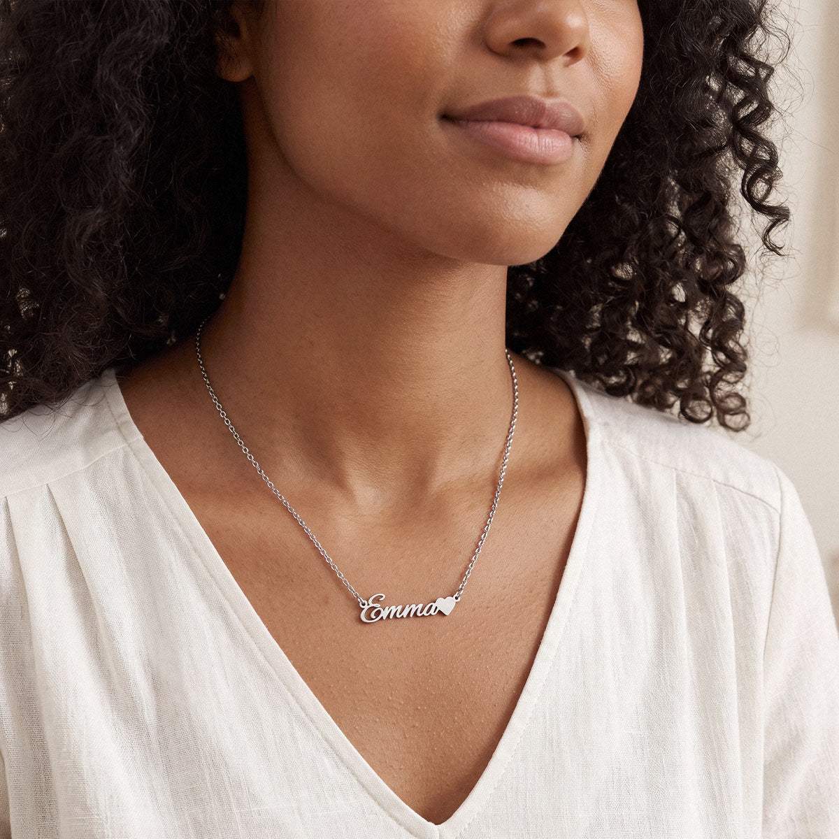 A close-up of a woman wearing a silver nameplate necklace that reads 'Emmanuel' over a white V-neck top.