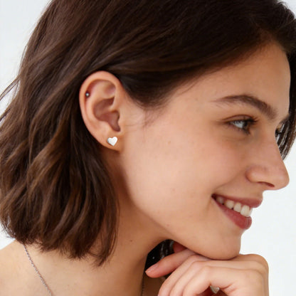 A close-up of a woman with short brown hair wearing a small silver heart stud earring on her lobe, shown in profile against a clean white background.