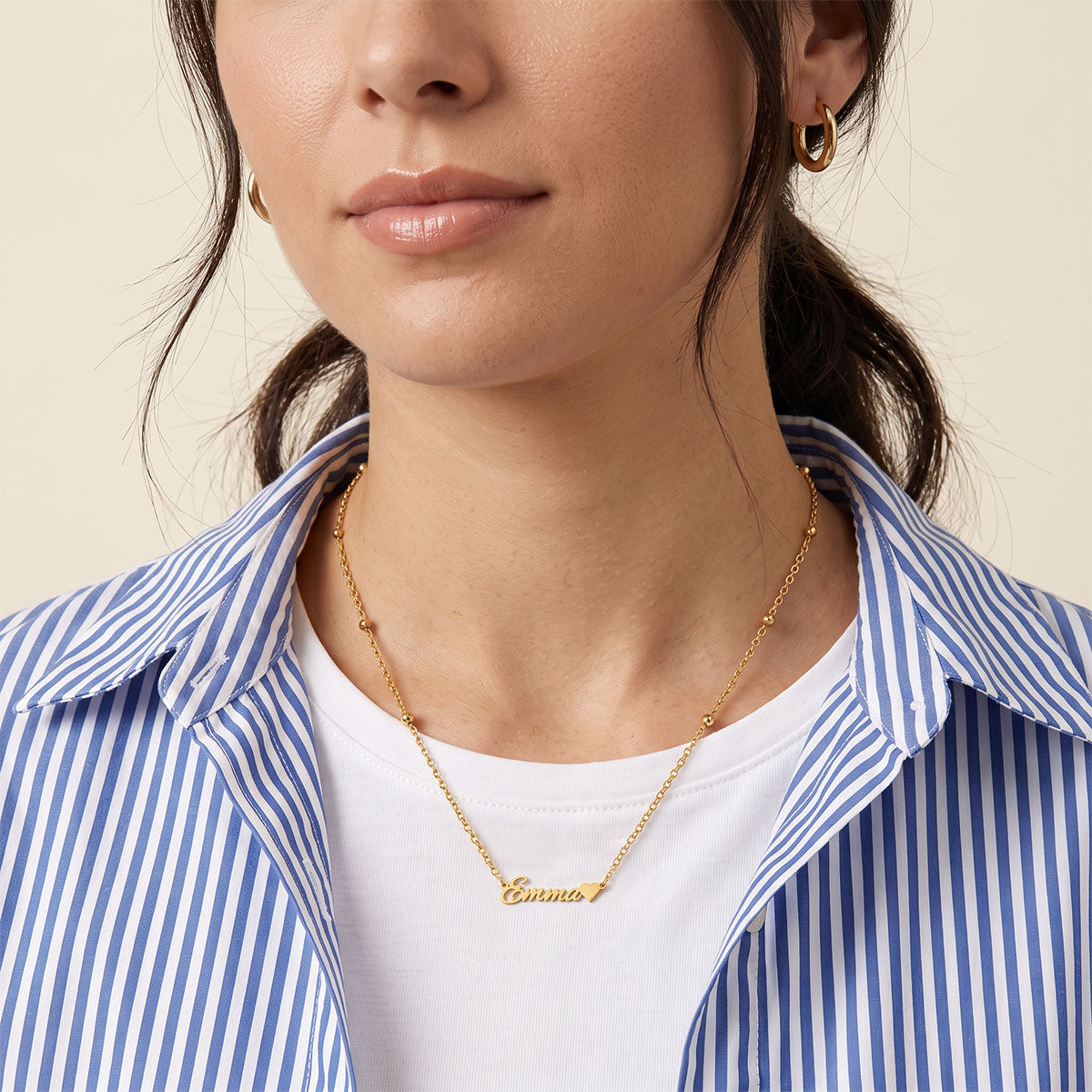 A close-up of a woman wearing a delicate silver nameplate necklace that reads 'Emma' on a fine chain, paired with a white V-neck blouse.