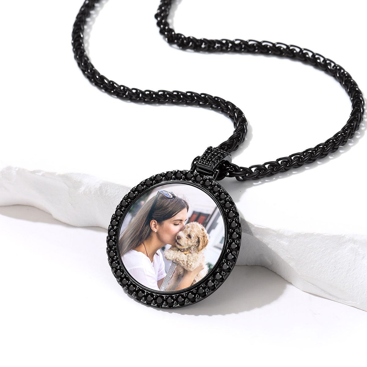 A custom photo pendant necklace on a black chain, featuring a circular black bezel set with small clear stones and a custom photo of a woman kissing a dog, displayed on a white textured surface.