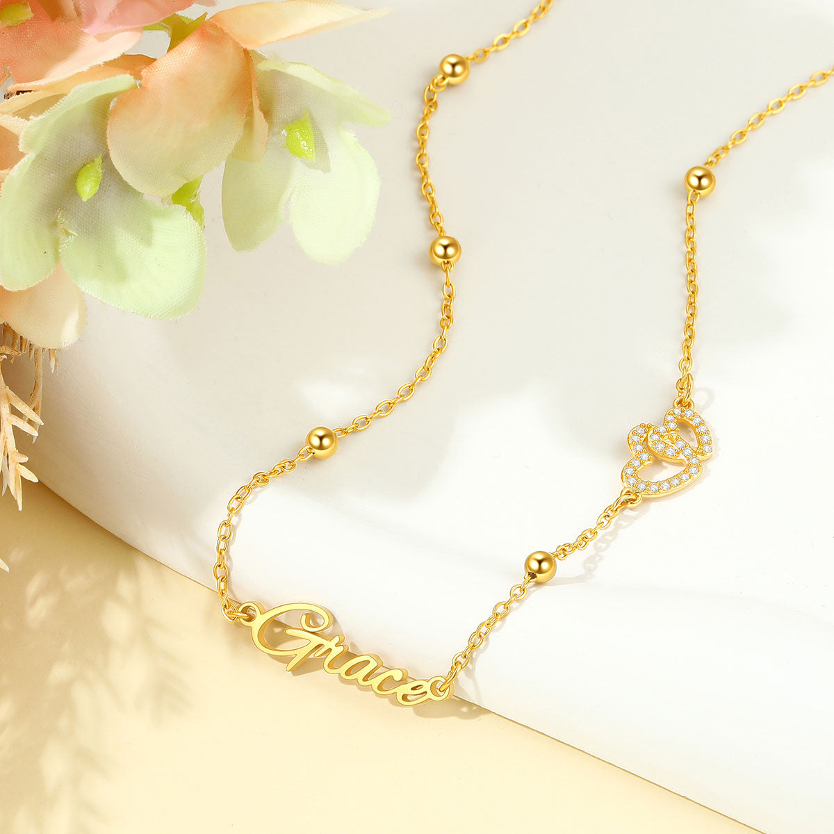 A delicate gold chain necklace with a script nameplate reading 'Grace' and a small crown charm embellished with tiny clear stones, styled on a white surface with soft peach flowers in the corner.