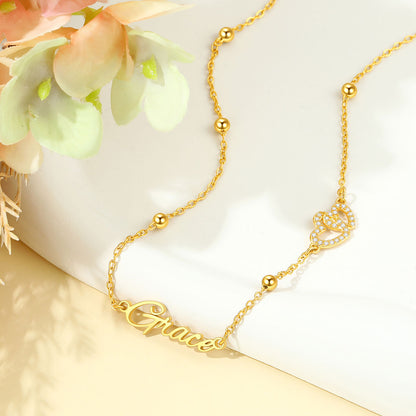 A delicate gold chain necklace with a script nameplate reading 'Grace' and a small crown charm embellished with tiny clear stones, styled on a white surface with soft peach flowers in the corner.