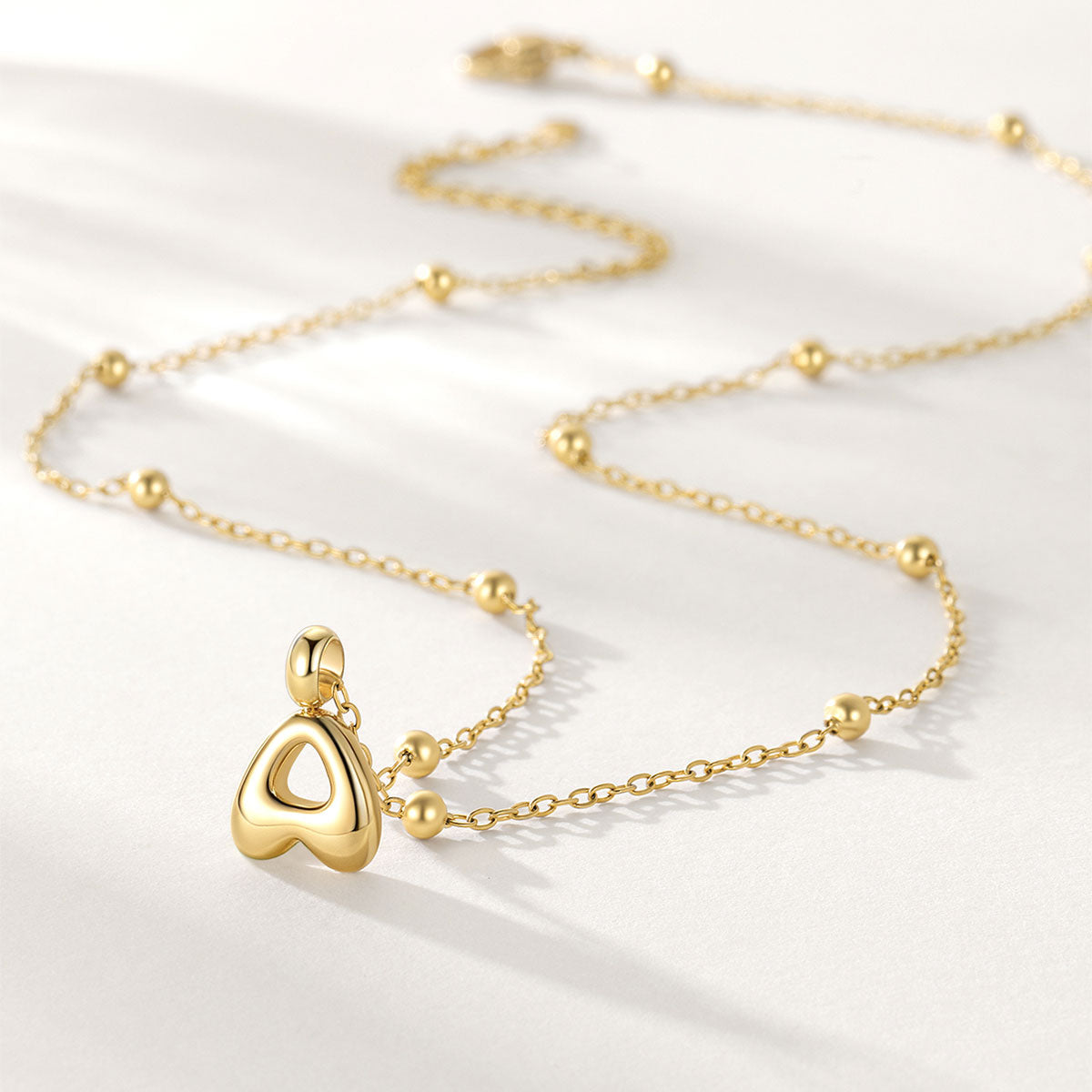 A delicate gold initial A necklace featuring a small, polished gold heart-shaped pendant with a cutout center, accented by small gold beads spaced along the chain, photographed on a clean white background.