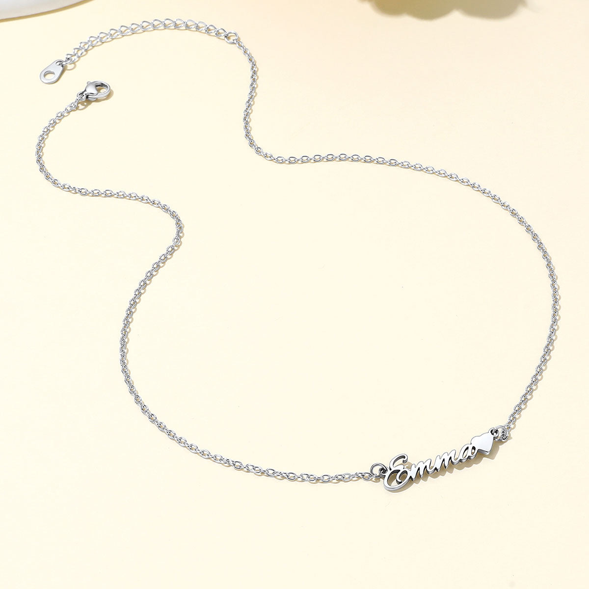 A delicate silver name necklace with a personalized script nameplate reading 'Emma' and a small heart, displayed on a light cream background.
