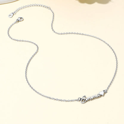 A delicate silver name necklace with a personalized script nameplate reading 'Emma' and a small heart, displayed on a light cream background.