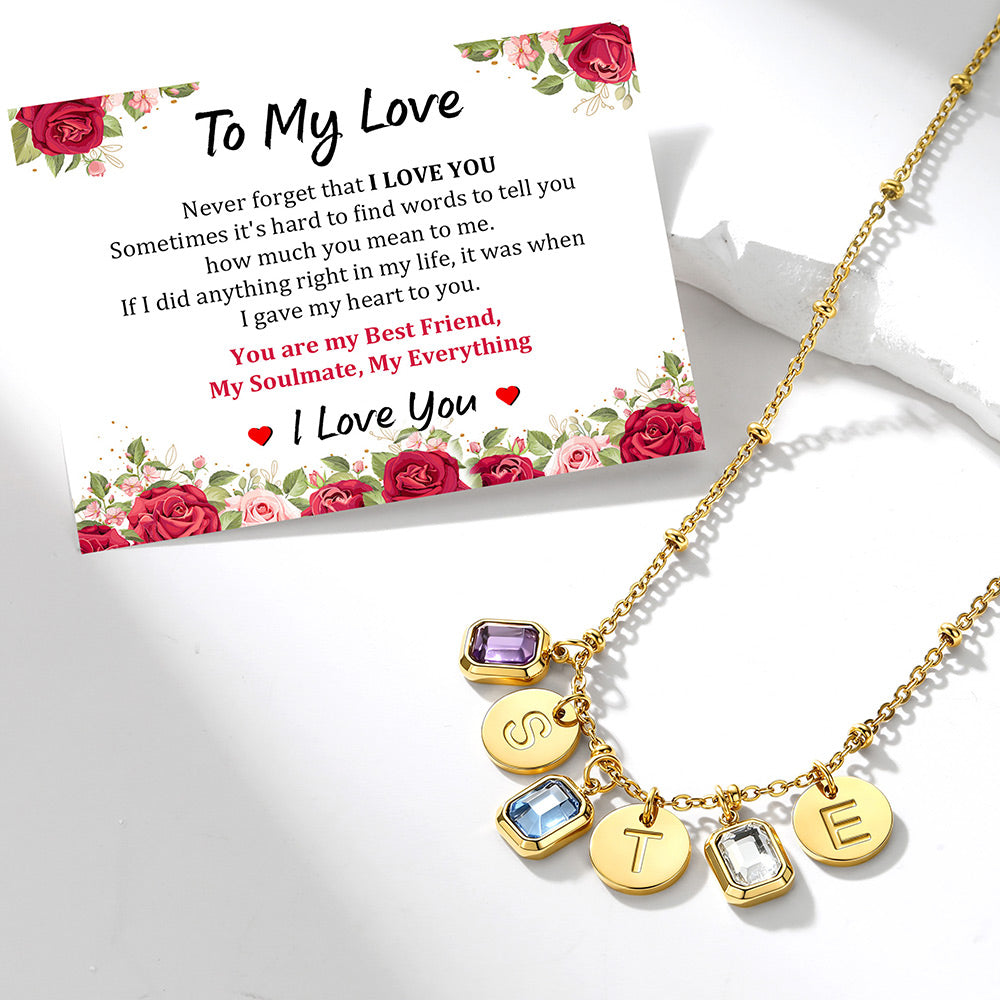 A gold-toned birthstone and initial charm necklace displayed on a white surface next to a printed message card. The necklace has a chain with five charms: a purple square gem, a blue square gem, a round gold disc engraved with 'S', a round gold disc engraved with 'T', and a clear square gem engraved with 'E'.