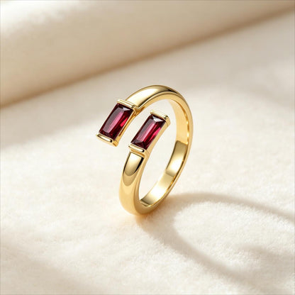 A gold birthstone ring with two rectangular dark red gemstones, displayed on a soft cream-colored fabric background.