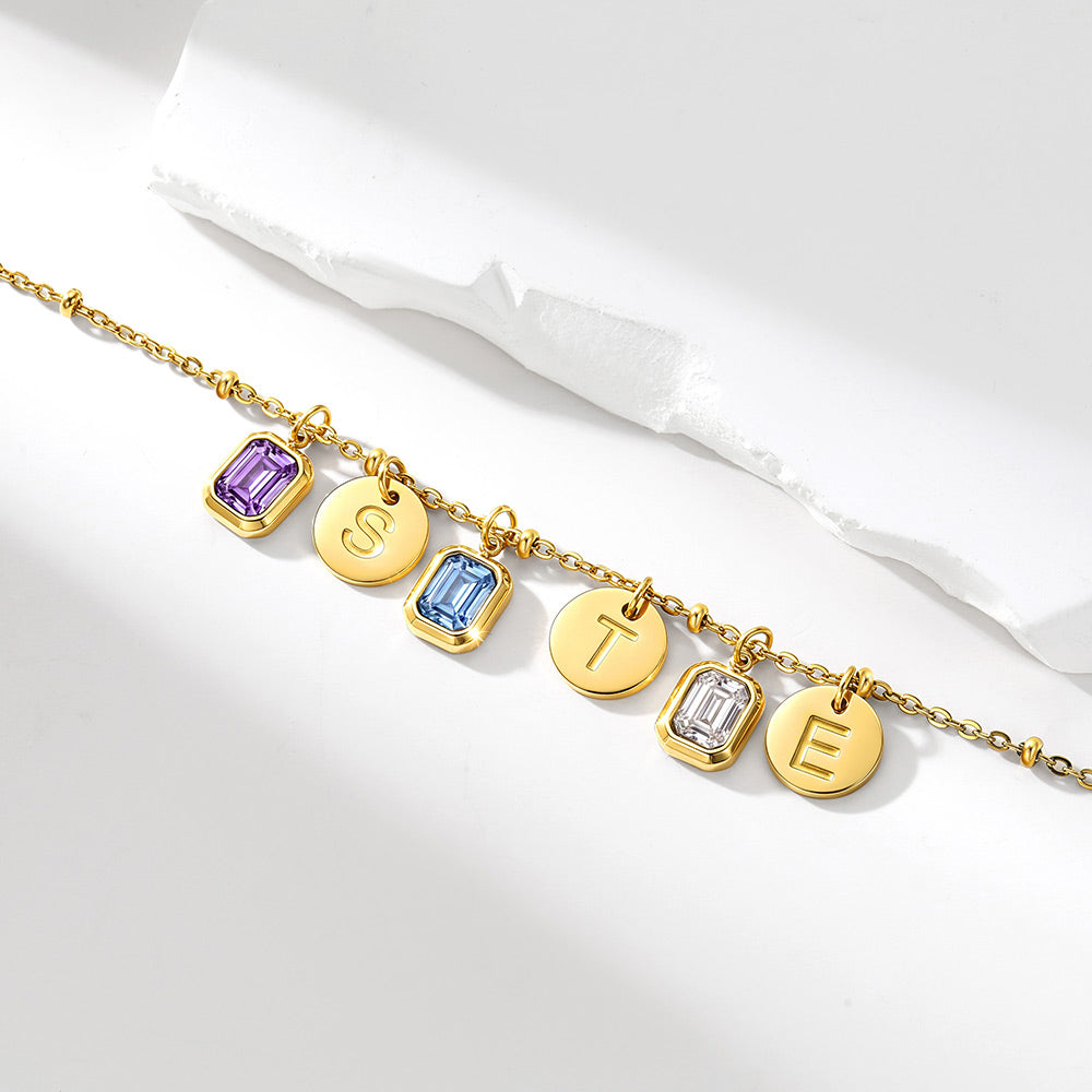 A gold chain birthstone necklace with five personalized charms: two round gold discs engraved with letters 'S' and 'T', and two rectangular gold bezel settings with clear gemstones in purple and light blue. The bracelet is displayed on a clean white surface with a textured white paper accent in the background.