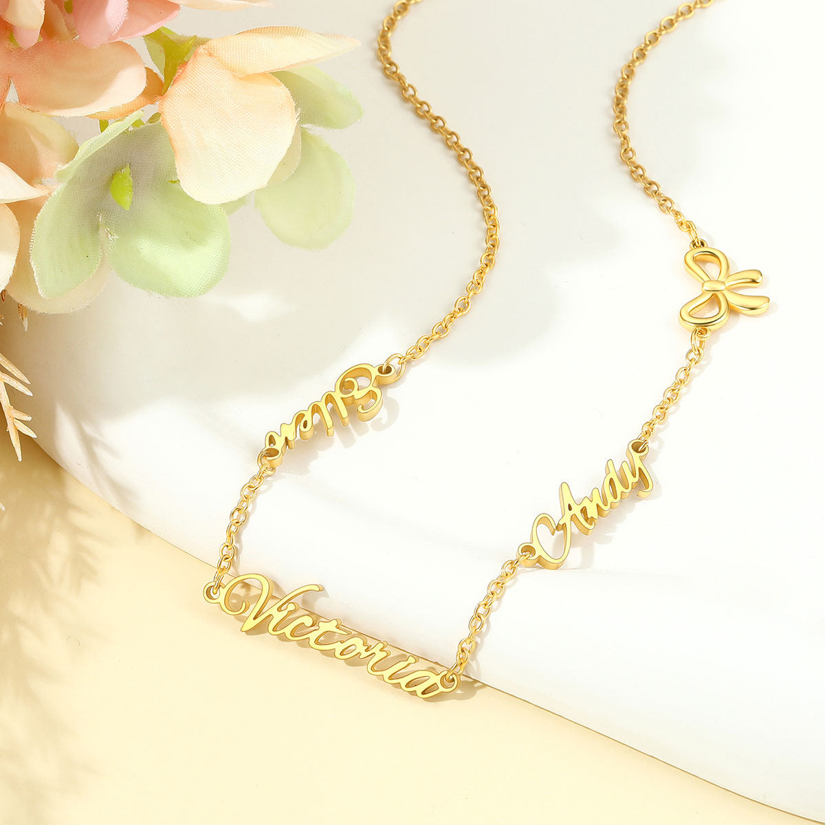 A gold chain name necklace with three personalized name plates and a four-leaf clover charm, displayed on a white surface with soft pink flowers in the corner.