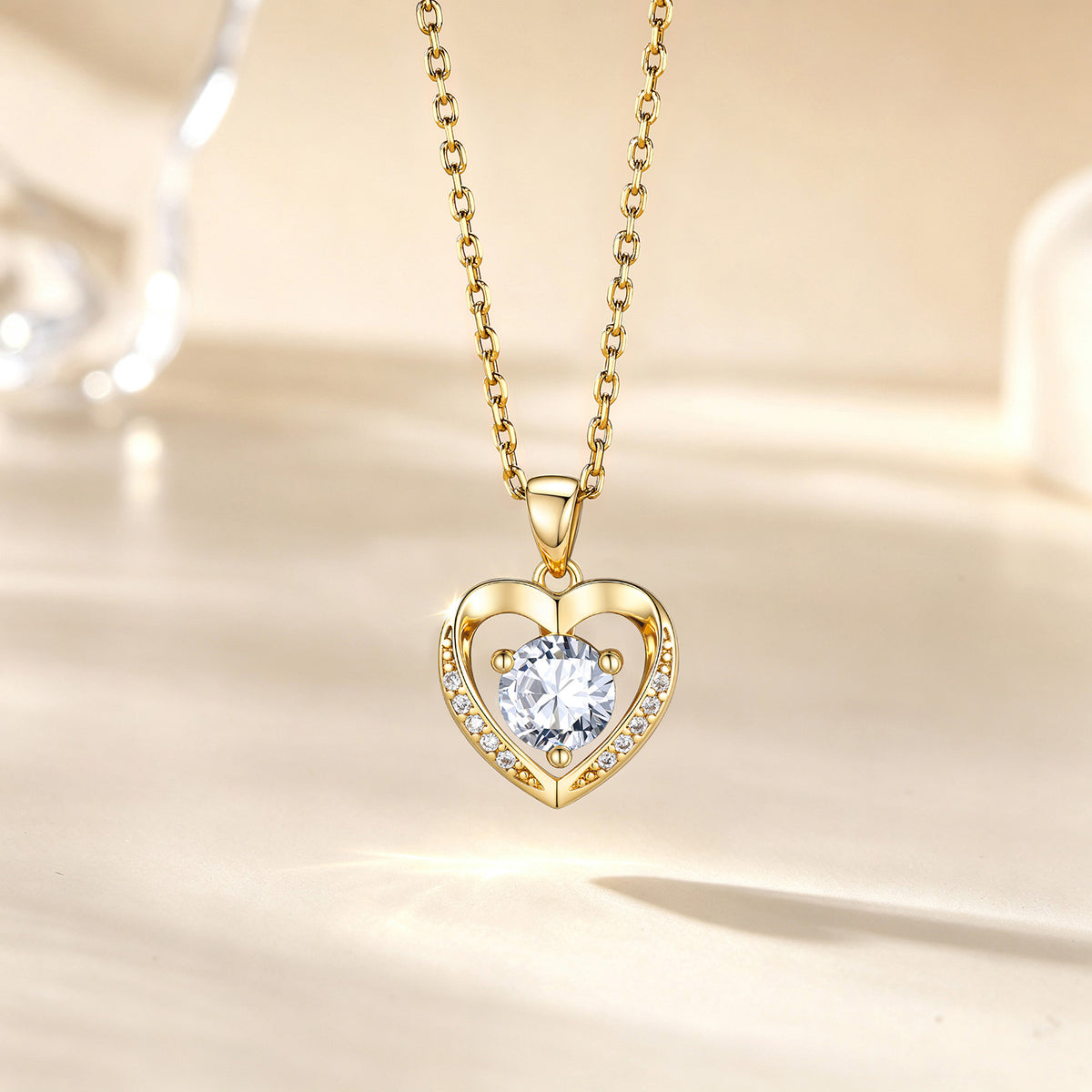 A gold heart pendant necklace with a clear round-cut center stone and small accent stones along the left side of the heart, hanging on a gold chain against a warm cream background.