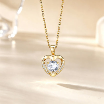 A gold heart pendant necklace with a clear round-cut center stone and small accent stones along the left side of the heart, hanging on a gold chain against a warm cream background.
