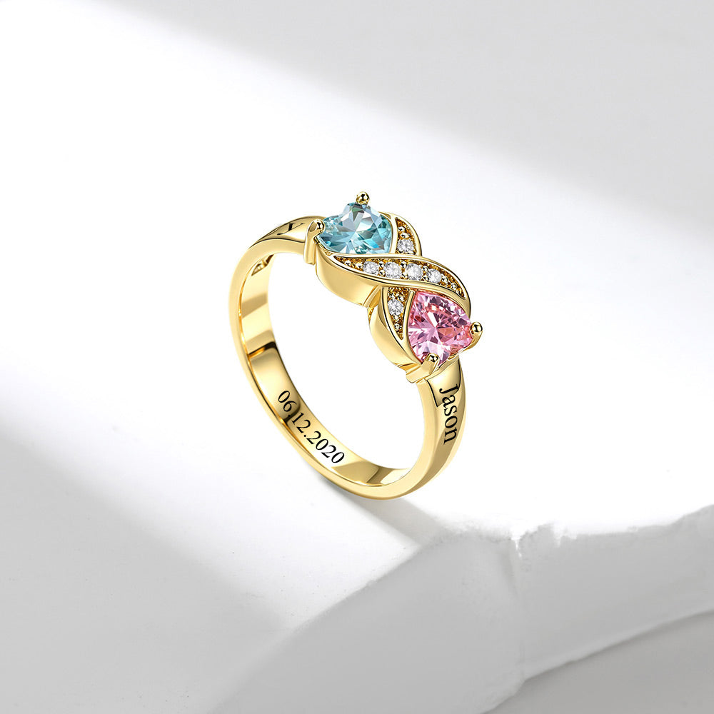 A gold infinity birthstone ring, featuring a light blue heart-shaped gemstone, a pink pear-shaped gemstone, and small clear accent stones on a white background.
