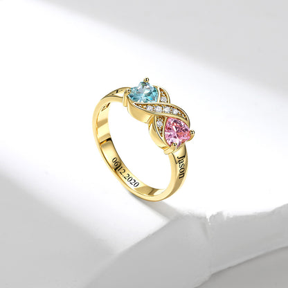 A gold infinity birthstone ring, featuring a light blue heart-shaped gemstone, a pink pear-shaped gemstone, and small clear accent stones on a white background.