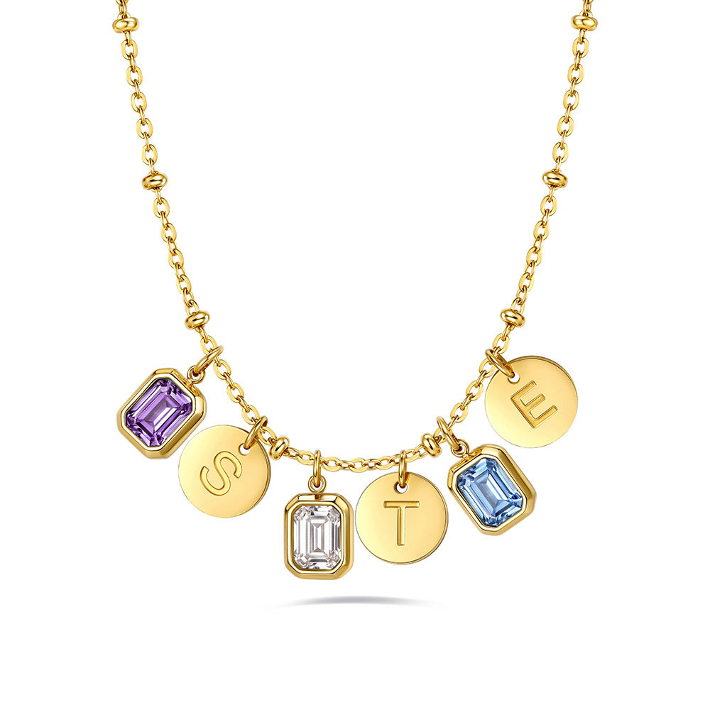 A gold initial birthstone necklace featuring five dangling charms spelling out 'STEL'. The charms include two round gold discs with engraved letters 'S' and 'T', one round gold disc with the letter 'E', one square bezel-set purple amethyst, and one square bezel-set light blue topaz, with one clear gemstone charm.