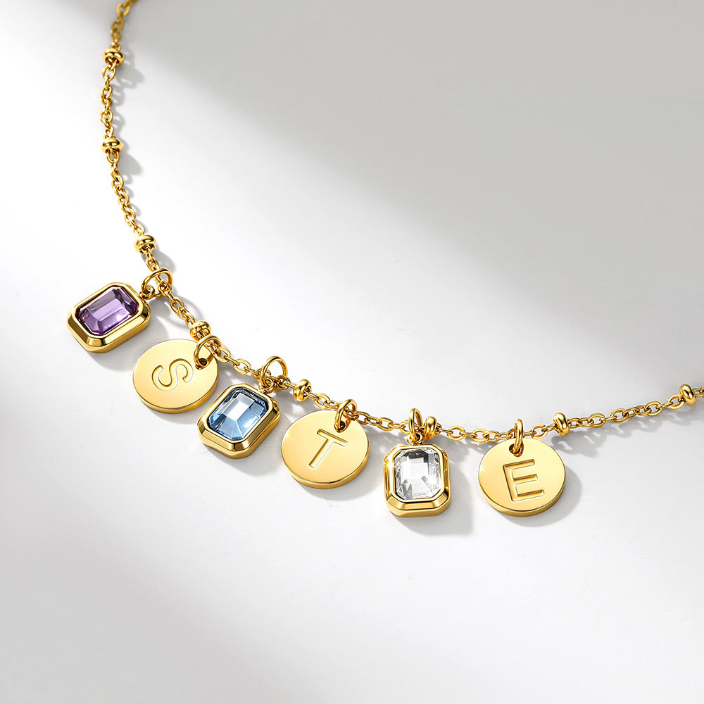A gold initial birthstone necklace featuring five round and rectangular charms set with purple, light blue, and clear gemstones, plus two engraved gold letters 'S' and 'E', displayed on a clean white background