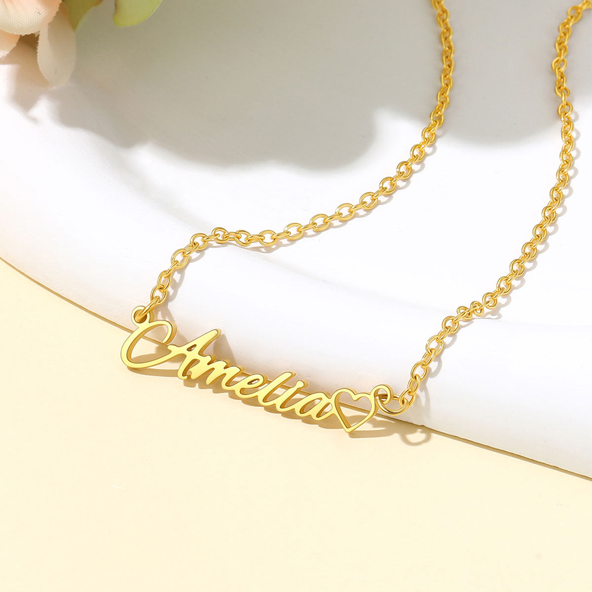 A gold name necklace with a personalized script nameplate reading 'Amelia' in cursive, with a small heart at the end, displayed on a white curved surface.