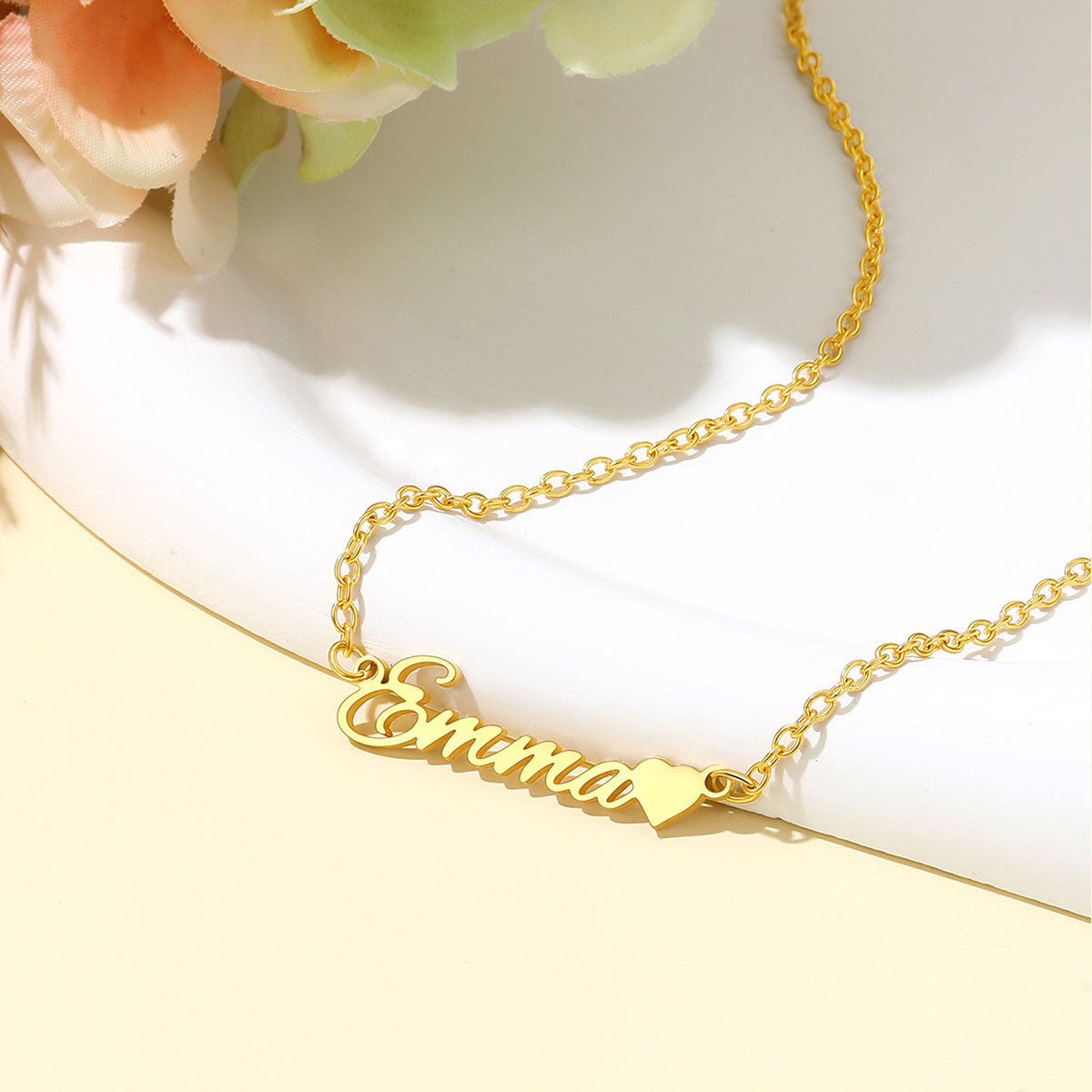A gold nameplate necklace spelling 'Emma' with a small heart charm, displayed on a white curved surface with soft peach flowers in the corner.