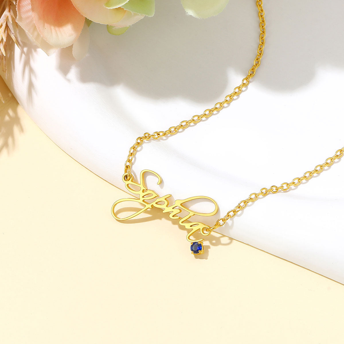 A gold nameplate necklace with the name 'Sophia' in cursive script, featuring a small blue gemstone accent, displayed on a white ceramic dish with soft peach flowers in the background.
