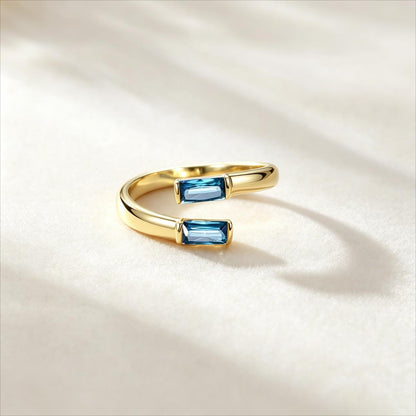 A gold open-ended birthstone ring featuring two rectangular blue gemstones, photographed on a soft white textured surface with elegant shadows.