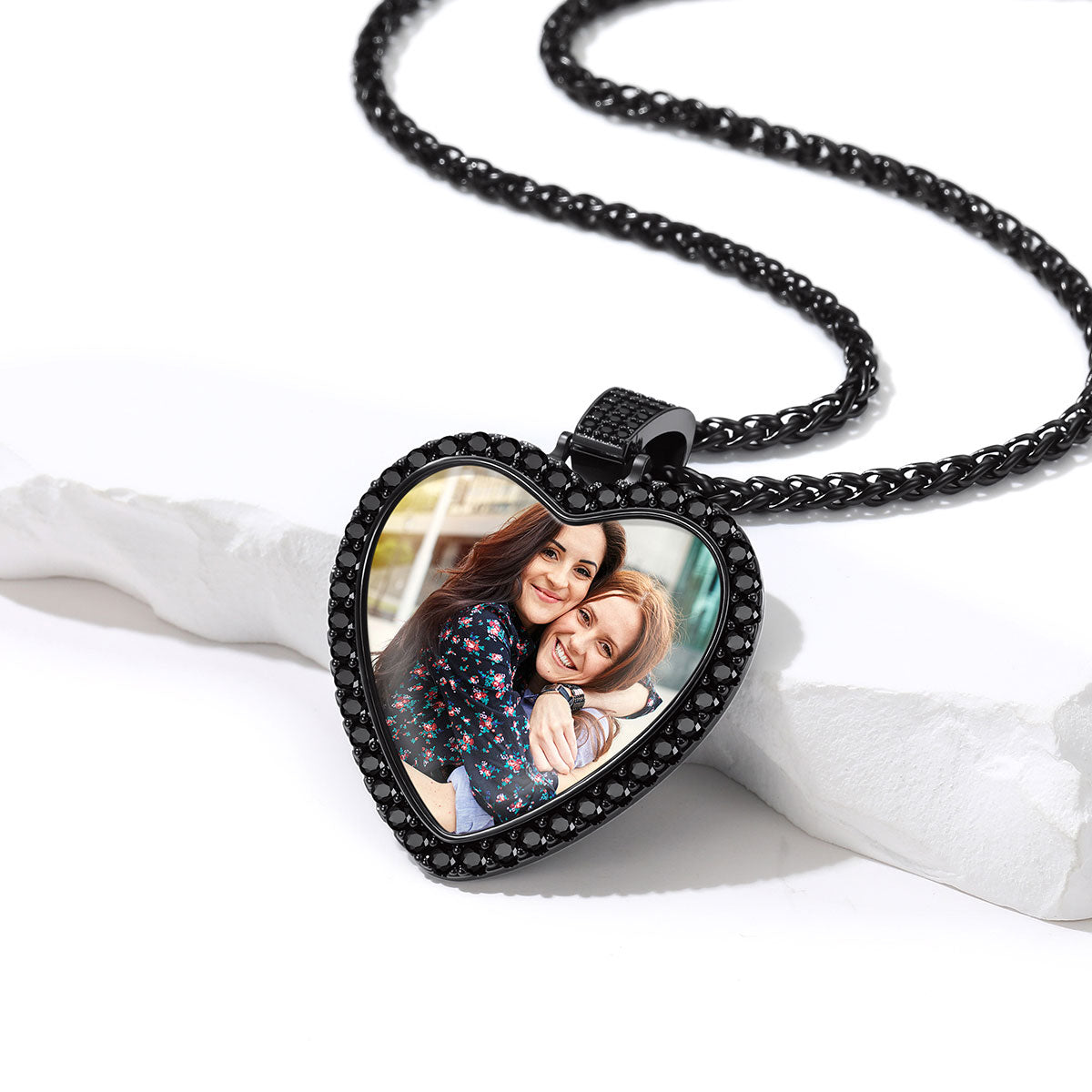 A heart-shaped pendant necklace with a black bezel set with small clear stones, featuring a custom photo insert of two women hugging. The necklace is displayed on a white textured stone prop against a clean white background.